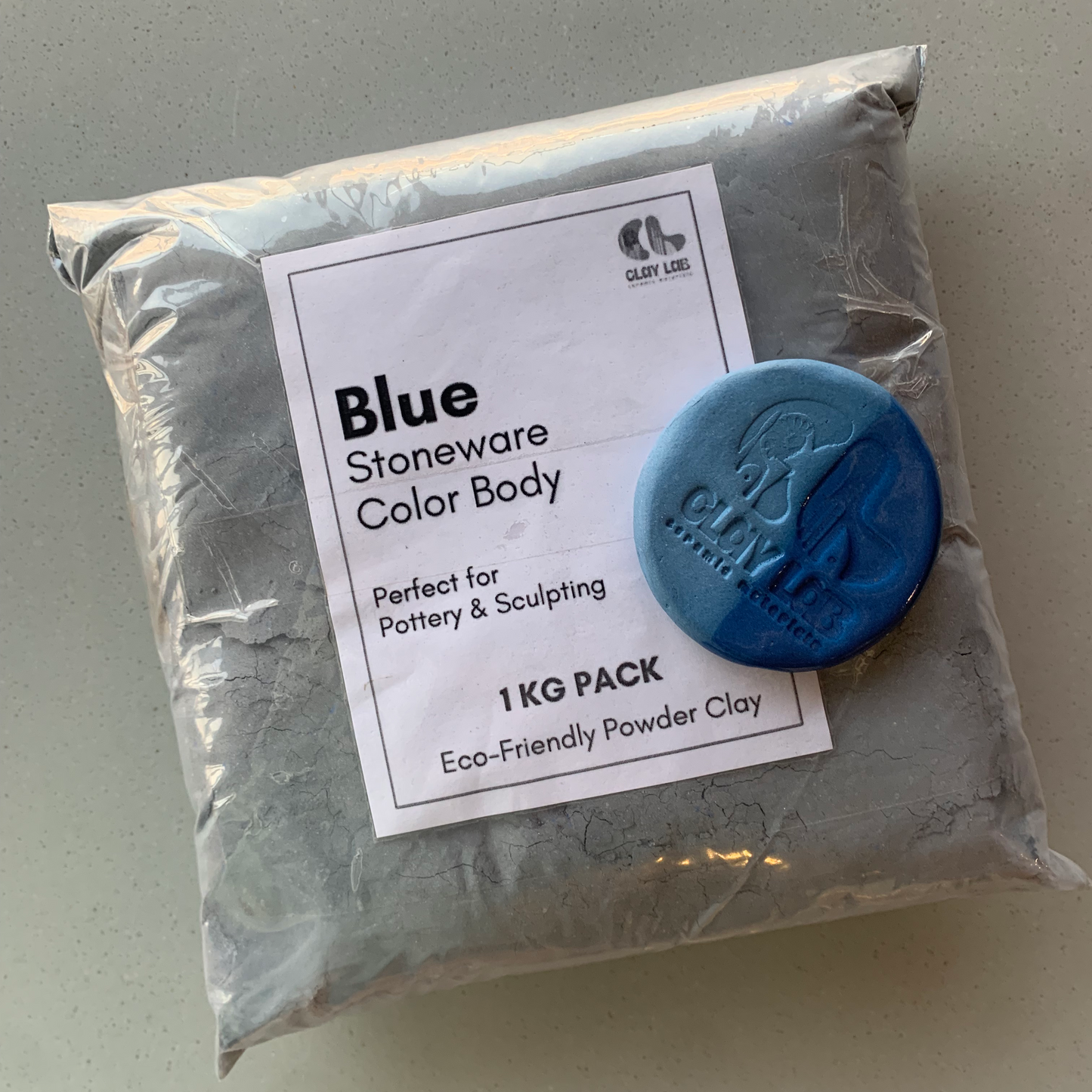 A sealed plastic bag containing blue stoneware color body powder clay with a label on top. A small sample clay piece is shown beside the bag, displaying the blue color.